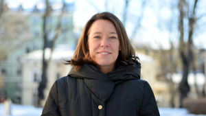 Åsa von Schoultz Professor i allmän statslära Åsa von Schoultz i vinterrock barhuvad på statsvetenskapliga fakultetens gård med ljusa byggnader och träd i bakgrunden.