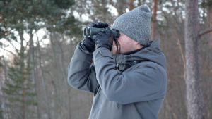 Populationsekolog och specialforskare Andreas Lindén. Man i grå jacka och mössa ser in i en kikare. Skog i bakgrunden.
