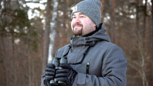 Ljuset styr flytten, berättar Andreas Lindén. Man i grå jacka och mössa med kikare i handen ler. I bakgrunden skog.