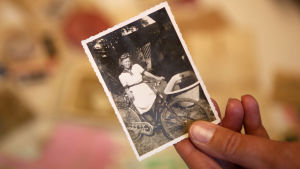 En person håller i ett gammalt svartvitt fotografi mellan tummen och pekfingret. På bilden står en ung flicka i vit klänning med en cykel.