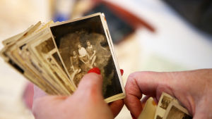 En person med röda fingernaglar håller i en bunt med mer än 20 gamla svartvita fotografier från 1940-talet. På den översta bilden sitter tre damer i klänningar i en glänta.