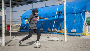 Ung kvinna i slöja spelar står vid ett fotbollsmål och sparkar en boll i ett flyktingläger. Ung kvinna i slöja spelar står vid ett fotbollsmål och sparkar en boll i ett flyktingläger.