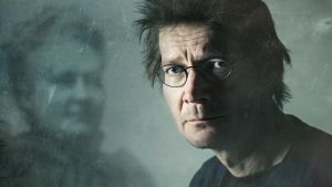 Minäkin olen Minna Canth. Juha Hurme katsoo kameraan taustallaan kuva, jossa näkyy sumea Minna Canth