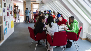 skolelever sitter och står i ett rum med fönster i snedtak. Röda stolar på blå heltäckande matta