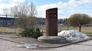 Sjundeå är i färd med att planera det nya skolcentret. Skylt där det står Aleksis Kiven koulu. Skylten är vid en gräsplan och grusväg.