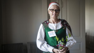 Läraren Ingelisa Wikholm hade tagit på sig en traditionell folkdräkt från Kyrkslätt för slava-festen. En kvinna står inne i ett hus. Hon har på sig en traditionell folkdräkt. I handen har hon en glasburk med en björkkvist och Serbiens flagga.