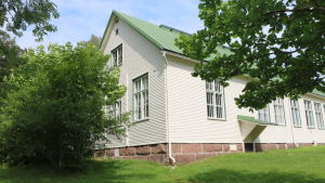 Rödskog skola är över hundra år gammal. Rödskog skolbyggnad. Ett vitt trähus. Det är sommar och gräset framför huset är grönt och träden brevid huset är fulla med löv.