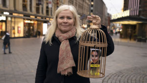 Latifas vän, finländskan Tiina Jauhiainen med en gyllene bur och en skylt där det står "Befria Latifa" Tiina Jauhiainen pitää käsissään kultaista häkkiä, jonka sisällä on prinsessa Latifa Al Maktoumin kuva.
