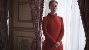 Prinsessan Anne, som hade ett svårt förhållande med medierna, spelas i The Crown av Erin Doherty. En ung kvinna i röd tröja står framför ett fönster med vita gardiner.