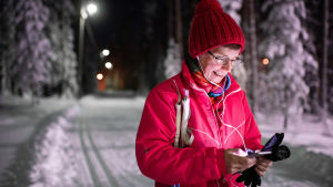 Keski-ikäinen nainen kuuntelee audiosisältöjä Yle Areenasta samalla kun on hiihtolenkillä talvisena iltana.