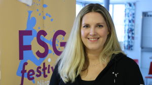 Hanna Stenholm efterlyser funktionärer och inkvartering. En ung kvinna med långt blont hår står intill en skylt med texten FSG-festival Närpes 2020.