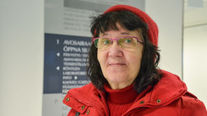 Eila Kipinoinen behöver besöka hälsocentralen ganska sällan. Hon har inte upplevt väntetiderna som alltför långa. Närbild på en leende kvinna i röd jacka och röd mössa.