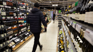 Många köpte rödvin till påskmiddagen. Alko uppmanade dock kunderna att hålla ett säkert avstånd till andra människor. Alkon myymälän sisällä pulloja hyllyissä. Miesasiakas tummansinisessä toppatakissa ja musta pipo päässä on juuri astunut kameran takaa kuvaan reippaasti kävellen hyllyjen välistä käytävää pitkin. Miehen liike on tallentunut epäterävänä.