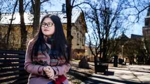 Qizai Wang i universitetsparken i Åbo. Asiatisk ung kvinna i glasögon sitter på parkbänk mellan trähus klädd i rosa kappa och grann halsduk.