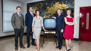 Sarjan hahmot seisovat isossa huoneessa vanhan television ympärillä.