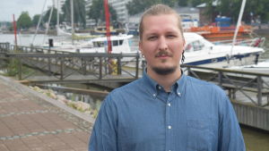 Ville Loukola berättar att han är nöjd med mängden material forskarna fått in.. En ung man i blå skjorta står framför en rad med båtar.