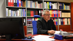 Man med grått hår och glasögon klädd i svart skjorta sitter framför en bokhylla fylld av pärmar.