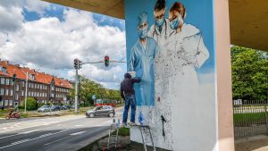 Varning för corona i Gdansk En man står under en bro och målar en graffiti på en bropelare av tre personer inom medicinska kåren och som alla tre ha munskydd.