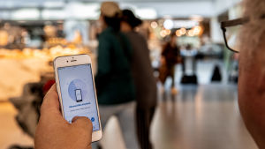 Coronablinkern bygger helt och hållet på frivillighet. Det är frivilligt att ladda ner appen och det är frivilligt att uppge om du fått ett positivt testresultat. En person har en mobil i handen och appen Coronablinkern är öppen och visas på skärmen. Bakgrunden med några människor och ljus är suddig.