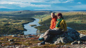 Nainen ja mies istuvat kivellä tunturin laella, taustalla tunturimaisema, jota halkoo joki.