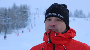 Janne Smeds En man med svart mössa och röd jacka står vid roten av en skidbacke.