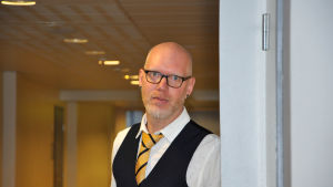 Kim Johansson är studiehandledare i S:t Olofsskolan. Kim Johansson i närbild, står lutad mot en vägg och tittar i kameran.