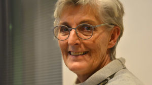 Margita Vainio. Margita Vainio, en glad dam med grått hår och glasögon med silverfärgade skalmar.