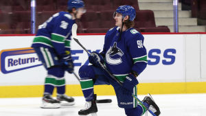 Vancouver Canucks spelare, här i form av Nils Höglander och Adam Gaudette, väntar ännu på nästa gemensamma träningspass. Klubbens coronasituation innebär att att NHL:s grundserie förlängs. Två blåklädda Vancouverspelare under uppvärmning, en tänjer på knä, en åker skridsko bakom.