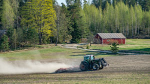 Vårbruket kan inledas ifall marken håller och vädret tillåter det. En grön traktor harvar jorden en vårdag där löven spricker ut i träden och en röd lada lyser i solen.