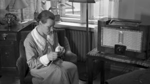 Nainen istuu kutomassa ja kuuntelee radiota. Mustavalkoinen kuva.