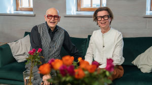 Jorma Uotinen ja Sami Sykkö istuvat sohvalla nauraen ja katsovat kameraan. Pöydällä heidän edessään on ruusuja maljakoissa.