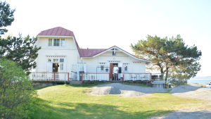 De Fyra Vindarnas Hus finns på Lilla Tallholmen i Hangö och besöks av många turister om somrarna. En gammal vit trävilla med rött tak vid havet, framför dörren står en man.