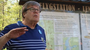 Calle Storm berättar engagerat om flygflottiljen i Jungfrusund. Calle Storm i blåvitrandig guidetröja framför en infotavla