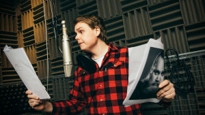 Itkisitkö onnesta? Mies lukemassa tekstiä mikrofoniin studiossa. Kädessä papereita ja Gösta Sundqvistin valokuva.
