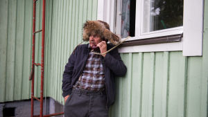 Heikki Kinnunen on pääroolissa elokuvassa Ilosia aikoja, Mielensäpahoittaja. Kverulanten står på gården och talar i trådtelefonen vars sladd hänger uyt genom köksfönstret..