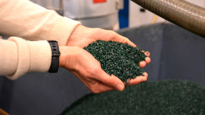 Granulat är små plastkulor som görs av återvunnen plast. Händer som håller i gröna plastpelletar ovanför ett stort fabrikskärl.