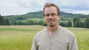 Tuomas Mattila har gedigen lista av titlar - han är utbildad diplomingenjör i kemi, magister i jord- och skogsbruk och teknologie doktor. En man står i en beige skjorta. I bakgrunden syns åkrar och skog.