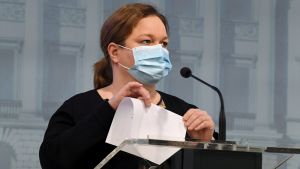 Den viljestarka omsorgsministern Krista Kiuru (SDP) ville inte kommentera frågan om obligatoriska gränstester. En kvinna i blått munskydd står vid en mikrofon. Hon är klädd i svart tröja och har håret i en hästsvans. I händerna har hon ett pappersark som hon viker på mitten.