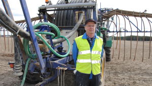 Timo Heikkilä driver en smågrisfarm i Rusko med 3 500 suggor. Grisarna säljs sedan vidare till andra gårdar för att gödas inför slakt. Man med arbetskläder står framför stor traktor på åker.