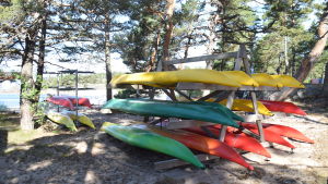 På Robinsonlägret kommer barnen att använda kajakerna som finns på området. Bilder på kajaker som är uppdragna på stranden. Kajakerna är uppsatta på en ställning av trä. Kajakerna är gula, gröna och röda. I bakgrunden syns havet.