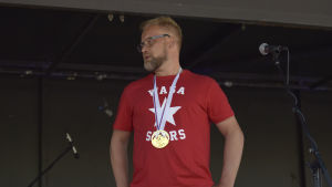 Mikko Manner. en skäggig man med röd t-skojrta och med en vm-guldmedalj runt halsen står på en scen