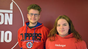 Kasperi Kaukonen och Julia Hämäläinen tycker att Kiviniemis föreläsning var givande. De anser båda att det är bra att kunna svenska i Finland. Två gymnasieelever i röda tröjor står bredvid varandra, framför en röd vägg.