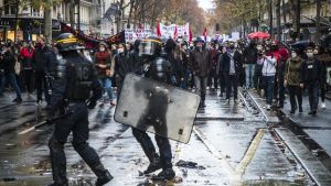 Under demonstrationer mot en föreslagen säkerhetslag i Frankrike, som den här i mitten av december, drabbades journalister av våld av både polis och demonstranter. Två poliser med hjälm sköld går på en våt gata framför en folkmassa med banderoller och paraplyer.