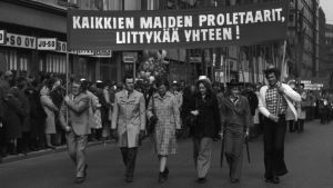 Förstamajtåg i Helsingfors på 1970-talet. Man visade färg, fast det var svartvitt. Arbetarnas valborgsmarsch 1972. Sex personer i förgrunden går med en banderoll.