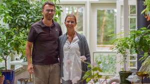 En glasveranda eller vinterträdgård som heter duga. Här får många blommor och grönsaker sin start på vårvintern. Ilkka Rissanen och Karin Björklund-Rissanen är väldigt nöjda över tillbyggnaden. En man och en kvinna inne i en glasveranda där det finns blommor.
