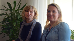Anne Ahtola-Poiesz och Maria Kronlund. två kvinnor sitter på stolar i en hall. I bakgrunden en grön växt. Solen lyser in från fönstret.