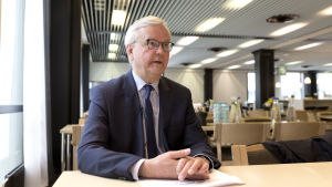 Professor Olli Mäenpää En äldre man i blå kostym sitter vid ett bord med händerna knäppta. Han ser allvarlig ut.