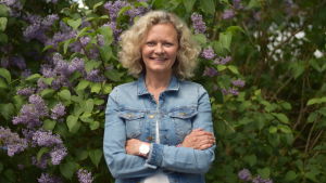 Turistsekreterare Martina Österberg arbetar med att marknadsföra Raseborg. En kvinna med blont lockigt hår och jeansjacka. Står framför en syrenbuske.