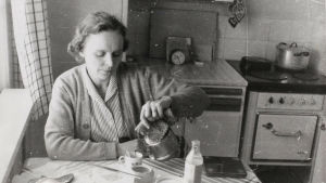 En kopp kaffe i eget kök. Sirkka-Liisa Miettinen i sitt hem. Svartvitt foto av Sirkka- Liisa Miettinen som häller upp kaffe åt sig själv sittande vid köksbordet.