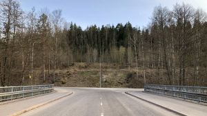 Samma område sett från bron över Svartån. Bostads- och affärsområdet breder ut sig uppe mittemot bron, både till höger och vänster sett från från bron. En vy från bron över Svartån mot Bruksvägen i Billnäs bruk. Mitt på bilden syns Banvallens motionsled. Asfalterad väg, skog, vår, bara lite grönska.